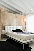 Schlafzimmer mit antiker Tischleuchte und Doppelbett mit weißem Leder vor Naturstein-Raumteilerwand