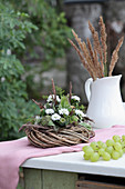Kranz mit weißen Astern und Mohnkapseln dekoriert und Krugvase mit Gräsern auf Gartentisch