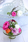 Colourful ranunculus on glass cake stand in front of white vintage container