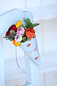 Colourful ranunculus in paper cone hung from backrest of chair