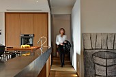 Open-plan, wooden designer kitchen with metal counter and woman in hallway