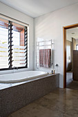 Badewanne mit brauner Mosaikverkleidung unterm Lamellenfenster