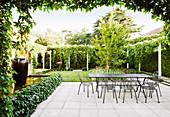 Terrassenplatz mit Tisch und Stühlen aus Metall, umgeben von grünen Pflanzen und Rasenfläche