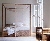 Four-poster bed with transparent curtains next to rattan bench and standard lamp