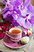 Tea in elegant teacup and saucer, green macaron and purple artificial flower