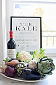 Teller mit Gemüse und einer Flasche Wein vor einem Bild am Fenster