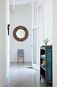 Blue chair and round mirror in bright hallway with glass wall