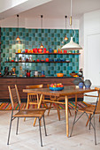Dining table and various chairs in front of kitchen with blue wall tiles