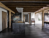Custom kitchen island made from black marble in open-plan interior with concrete floor and rustic wooden ceiling beams