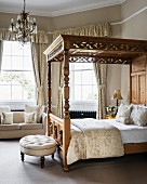 Ornate four-poster bed with scatter cushions in bedroom with bay window and buttoned-tufted stool