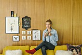 Young woman sitting on backrest of sofa next to small gallery of pictures