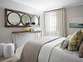 Mirror with wooden frame above console table and white ceramic drum stool at foot of double bed in bedroom
