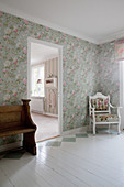 Old bench and upholstered chair flanking doorway in interior with floral wallpaper and painted floor