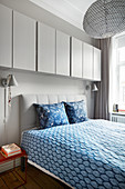 Grey, wall-mounted cupboards above bed with blue-patterned bed linen