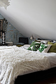 White furry blanket and leaf-patterned scatter cushions on bed below sloping ceiling
