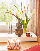 A statue of Buddha and gladioli on a pedestal in front of a window