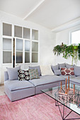 Grey corner sofa with scatter cushions, coffee table with glass top and plants on windowsill in front of interior doors with glass panels