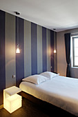 Double bed against wallpaper with broad dark stripes, pendant lamp and illuminated bedside cabinet in bedroom
