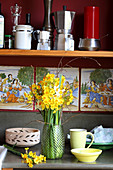 Narcissus, cream narcissus and birch twigs in vase, picture tiles and kitchen utensils on shelf