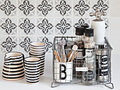 Spices in wire basket next to striped beakers and jug against tiled splashback