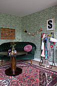 Round wooden table, velvet sofa and clothes rail against green wallpaper