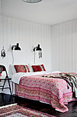 Floral cover on double bed below black reading lamps mounted on white wood-clad bedroom wall