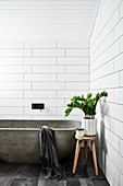 Concrete bathtub and houseplant on stool in white-tiled bathroom
