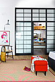 Roter Polsterhocker auf gemustertem Teppich, Stuhl, Poster und Wandlampe im Schlafzimmer neben geöffneter Badtür