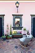 Rocking chair, foliage plants, bench and mirrored window with lava stone frame on pink wall of courtyard