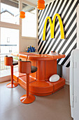 Orangefarbene Theke mit Barhockern vor schwarz-weiß gestreifter Wand mit Deko-Buchstabe