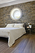 Double bed below porthole window in bedroom with stone wall