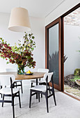 Runder Tisch mit Blätterzweigen und weiße Stühle vor offener Terrassenschiebetür