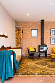 Log burner, retro armchair, stacked firewood and sofa in open-plan interior with brick wall
