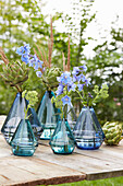 Blue glass vases with delphiniums and artichokes on a wooden table in the garden