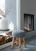 Stool with crocheted cover in front of stacked firewood and fireplace