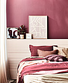 Bed with cushion, above it decoration on wall ledge in bedroom in shades of bordeaux