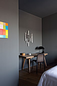 Paper artwork on wall above wooden table with black table lamp and chair in bedroom with grey walls