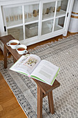 Book and teacups on wooden bench next to white glass-fronted cabinet