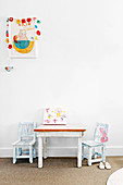 Children's table with two chairs in front of white wall, above with picture with garland