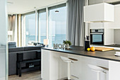 Raumhohe Fensterfronten mit Blick aufs Meer daneben offene Küche mit Küchentheke und Barhockern