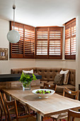 Chairs around wooden table in front of corner bench in bay window with shutters