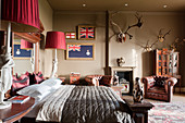 Grand mahogany bed with mirrored headboard in bedroom with leather buttoned armchairs, framed flags and mounted antlers