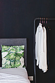 Kissen auf Bett mit grauem Bettkopfteil, daneben Kleiderstange vor schwarzer Wand