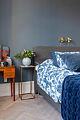 Dressing table and vase of flowers on slimline table next to grey box spring bed with blue-and-white bed linen