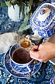 Tee wird aus blau-weißer Teekanne durch antikes Teesieb in Teetasse gegossen