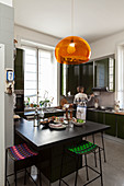 Pendant lamp with orange lampshade above counter and barstools in kitchen and woman in background