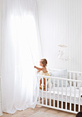 Weißes Babyzimmer mit Gitterbett und deckenhohen transparenten Vorhängen
