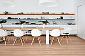 Long white kitchen counter below wooden shelves and dining table with shell chairs