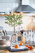Fir branch in swing-top bottle. glasses and cutlery on tray behind tangerines on folded tea towels