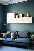 Blue sofa with scatter cushions below white wall-mounted cabinets on blue living room wall